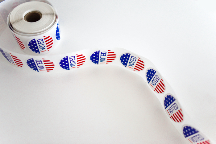 A roll of stickers is on a white table. The stickers are on their sides. Each sticker has a blue area on top, with small white stars in it. The middle of the stickers say "I voted". The bottom part of the sticker has horizontal red and white lines.