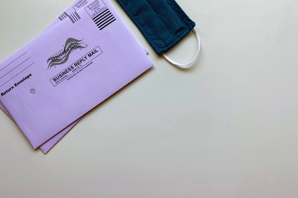 Two purple envelopes that say "Official Election Mail" on them next to a dark blue mask.