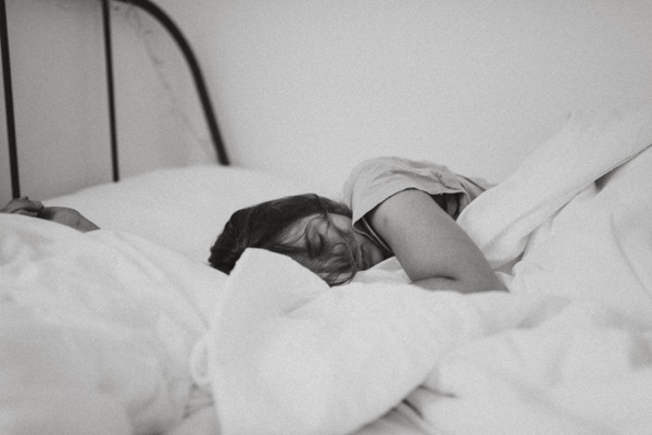 A grayscale photo of a sleeping woman. She is wearing a t-shirt and is using white blankets and pillows. The headboard is metal bars. Photo by Kinga Cichewicz on Unsplash
