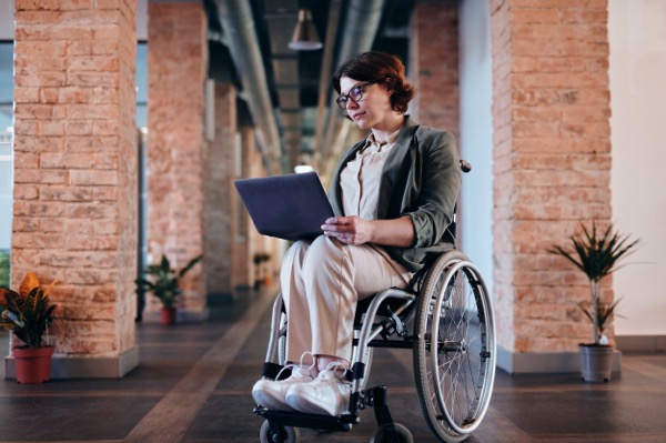 Photo of a woman looking at a laptop. She is using a wheelchair. Photo by Marcus Aurelius from Pexels.