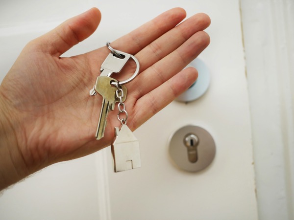 A person holds a set of keys with a keychain that looks like a house. Behind the hand is a slightly blurry door to a home. Photo by Maria Ziegler on Unsplash