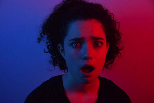 Woman with a shocked expression by Mason Kimbarovsky on Unsplash. Half of the woman's face is in red light, the other half is in blue light. She has curly dark hair.