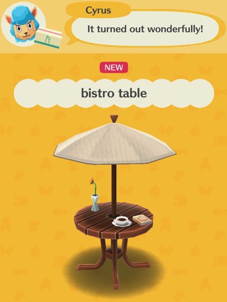 A bistro table has a tan umbrella covering it and providing shade. The umbrella is on a pole that goes through the wooden table below it. The table has four wooden legs. There is a flower in a small vase, a cup of coffee, and a book on the table.