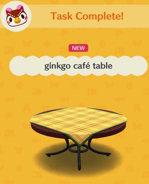 The gingko café table is a round wooden table with a metal base. The legs of the table are metal. There is a yellow and brown checkered tablecloth on the table top.