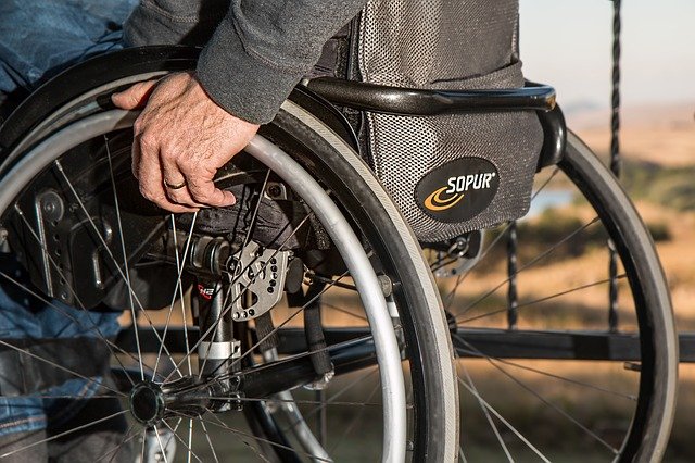 Photo of an older person using a wheelchair. The photo focuses on the back of the wheelchair, which has room for a backpack. The wheels of the chair are the most prominent part of the photo. Photo by Steve Buissine from Pixabay.