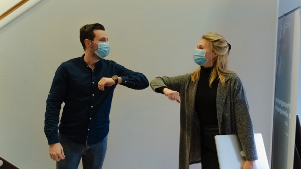 Two people are wearing masks indoors. They are doing an elbow bump. Photo by Maxime on Unsplash.