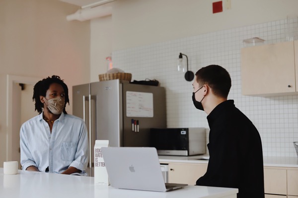 A black man in a light blue button down shirt is wearing a tan face mask. A white man in a black shirt is wearing a black face mask. They are in an office environment. Photo by Good Faces on Unsplash.
