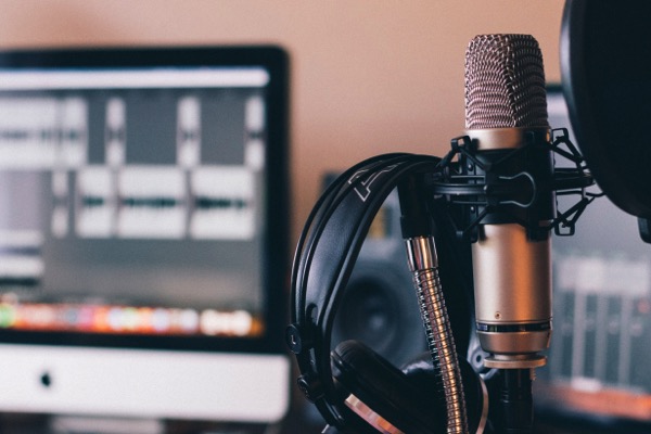 A microphone with a pop filter is next to a headset. In the background is a computer monitor with audio edition software on the screen. Photo by Will Frances on Unsplash.