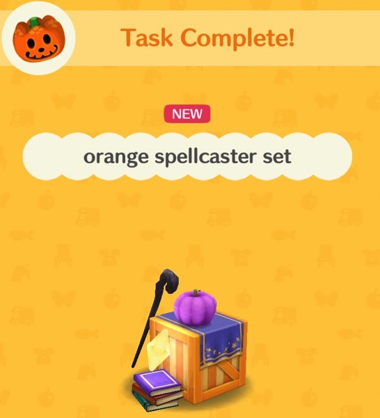 The orange spellcaster set has an orange and yellow box made of wooden slats. On top is a purple cloth  with gold lines around the edges and some gold stars on one side. A crooked staff leans on the box. A small purple pumpkin sits on top of the box. A small stack of books is on the floor, with a yellow star on top of it.
