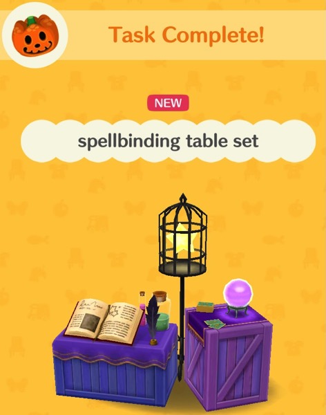 The spellbinding table set includes two purple wooden boxes. One has a purple orb that is sitting on a stand. There are cards next to it. The other has an open book, an inkwell, and some tall glass jars with corks in the top. Each jar has a different colored liquid inside it. Between the boxes is a tall birdcage on a stand. A yellow star is floating inside the cage.
