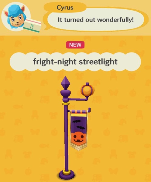 A purple post holds up a round streetlight that looks like an orange pumpkin. There is a banner hanging off the streetlight that is purple, with two black bat shapes flying on it. Below the bats is an orange pumpkin that has pointed ears and bat fangs.