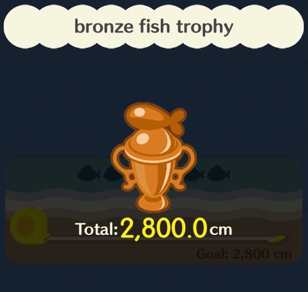The bronze fish trophy is much shinier than the wooden fish trophy. It is shaped like a standard trophy, with two jug handles on each side. There is a round, stylized fish on the top.
