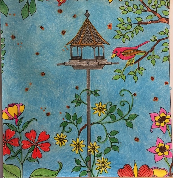 A bird feeder on a tall post is in the center of the coloring page. A red bird, that is stand on a branch that has leaves on it, looks at the feeder. A variety of flowers are growing at the bottom of the page. There appears to be a lot of pollen in the air.