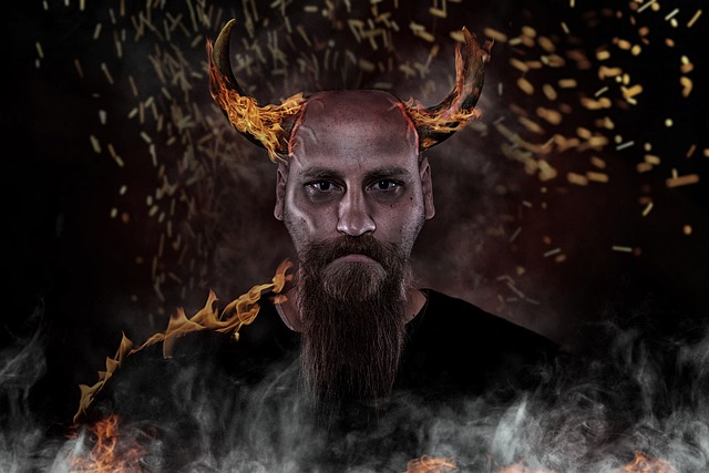 A man with a long beard looks at the viewer. The man has an intense look, and there are horns on his head. The horns have fire flowing over them. The man is wearing a black shirt that has fire running down one side, and a lot of smoke beneath him. Small bits of fire are floating in the air behind him. Photo by marla66 on Pixabay.
