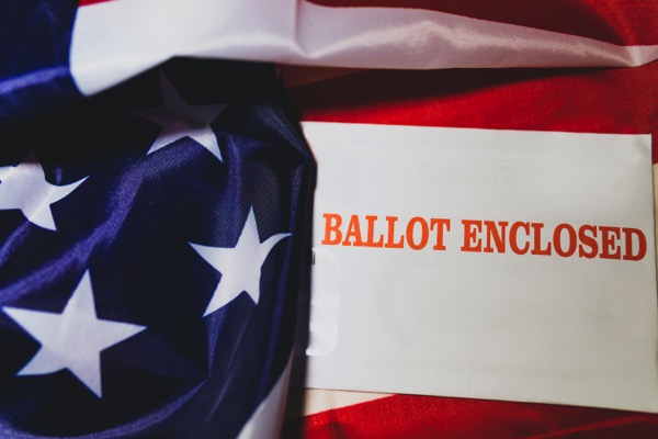 A sign says "Ballot Enclosed". It is on top of the American flag. Photo by Joshua Woroniecki on Unsplash