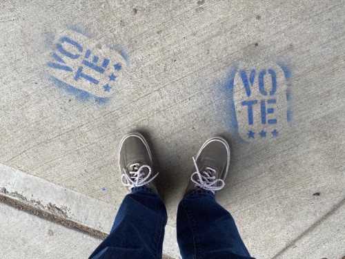 Person wearing blue pants and grey shows stands between two stencil on the sidewalk that say "VOTE"