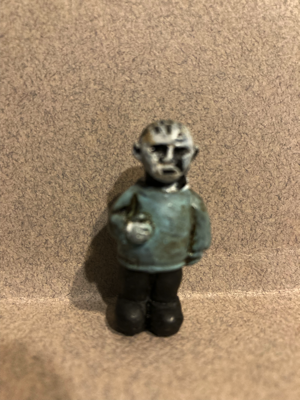 A hard plastic figure has silver skin. There are small holes on the top of his head that are where the pins would go. He is wearing a green sweater, black pants, and black boots. This figure is Pinhead from the Hellraiser movies.