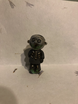 A hard plastic figure with a grey face resembles the character "Barrel" in "The Nightmare Before Christmas" movie. He is wearing a black shirt with bones on it, and black pants with bones on it. For some reason, his feet are green.