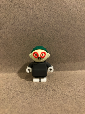 Day Six gave me this little guy. He has green hair and is wearing red circles around his eyes, and appears to have a small black mustache. His clothing is black. His face is a light yellow color. His hands look like LEGO hands and his feet look like LEGO feet.