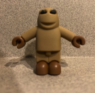 A brown plastic figure with a light brown head and body and dark brown hands and shoes. This one has a soft texture to it, and allows you to gently bend its arms. The face of this figure has black eyes and a smiling mouth. The hands look like something a Lego character would have.