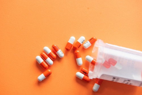 A clear plastic bottle is spilling out orange and white pills onto an orange table. By Christina Victoria on Unsplash