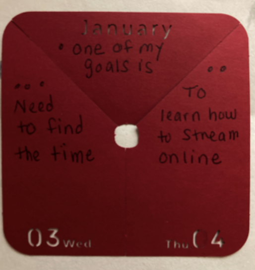 A red piece of paper with a hole in the middle has a haiku written on it: One of my goals is / to learn how to stream online / Need to find the time