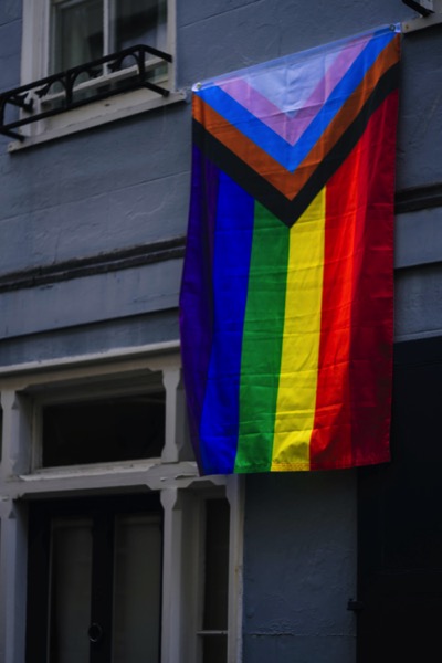 A flag with the transgender colors, a brown stripe and a black stripe, and the rainbow flag hangs on a house. Photo by Jack Lucas Smith on Unsplash