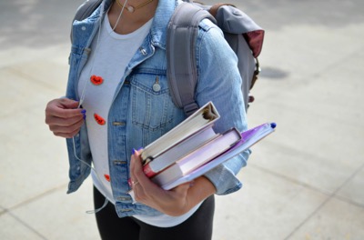 woman wearing a denim jacket with and carrying a backpack. She holds a stack of books in one hand - Unsplash